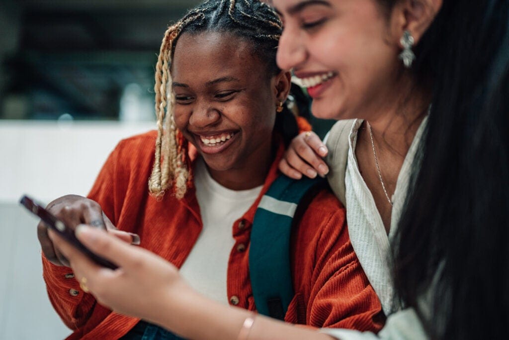 Two young women smiling and laughing while looking at a smartphone together, one with braided hair and a red jacket, the other with long hair and earrings.